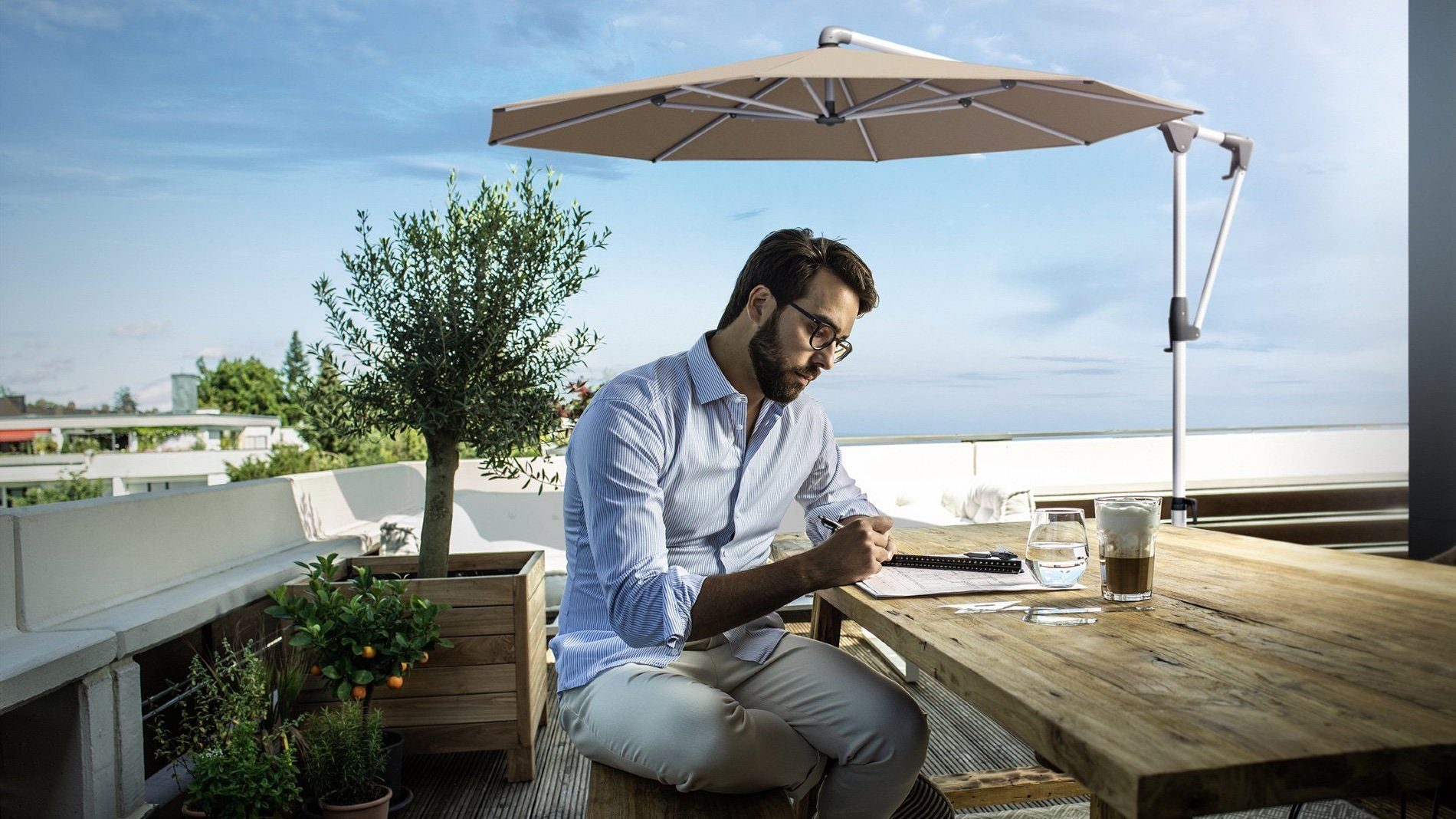 Mann sitzt an einem Holztisch auf einer Terrasse, Ampelschirm im Hintergrund