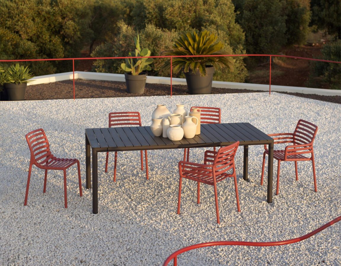 Ein Tisch mit fünf Gartenstühlen auf einer großen Terrasse mit Kies, im Hintergrund Pflanzen.